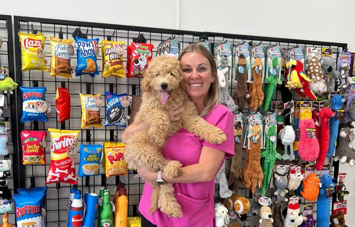 A woman in a pink shirt holding a curly-coated golden puppy beside a display of dog toys