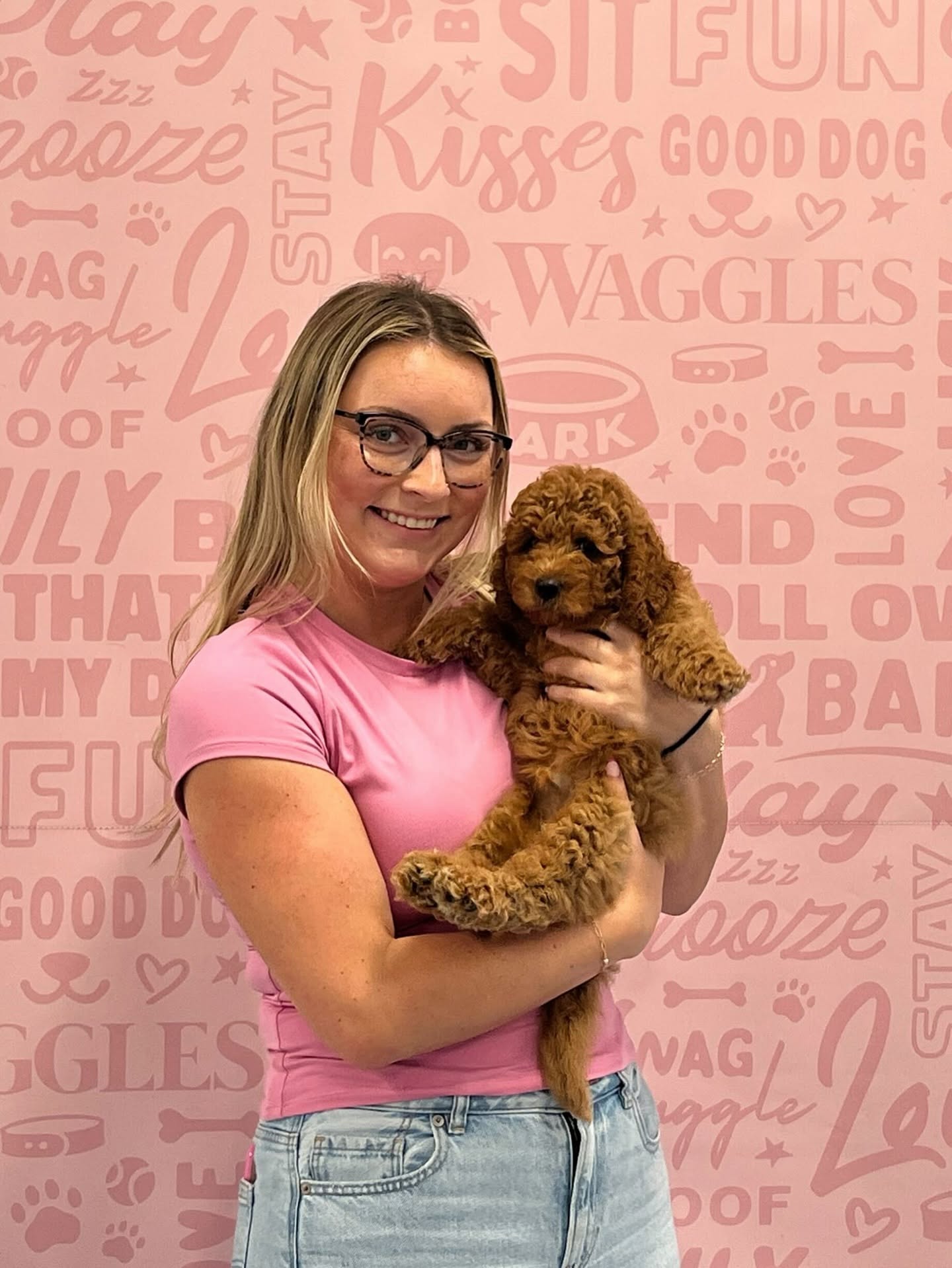 A woman holding a small brown curly-coated puppy in front of a pink Waggles wall